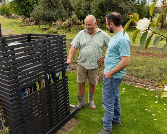 Two men standing next to a black compost bin in a garden.